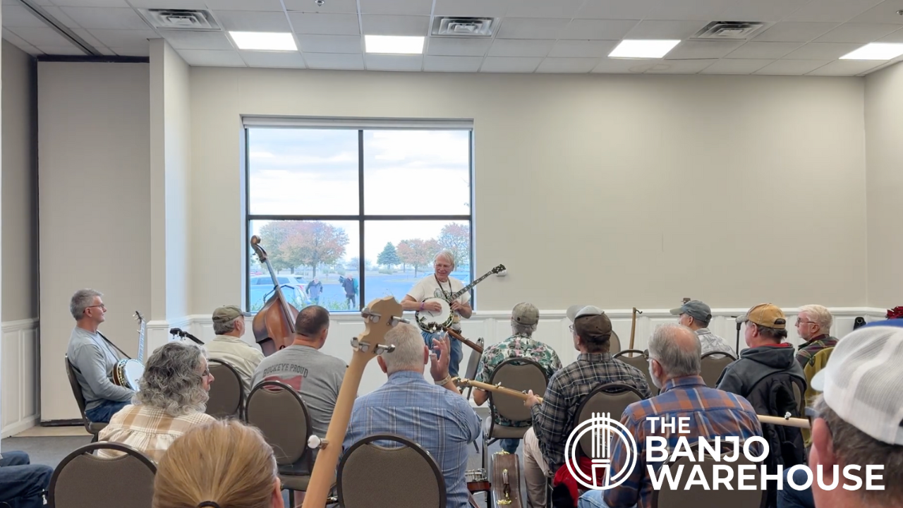 Geoff Hohwald stands at the front of a workshop room playing banjo for a seated audience of musicians. Attendees hold banjos and other instruments including an upright bass. Large windows behind him show fall foliage outside. The Banjo Warehouse logo appears in the bottom right corner.