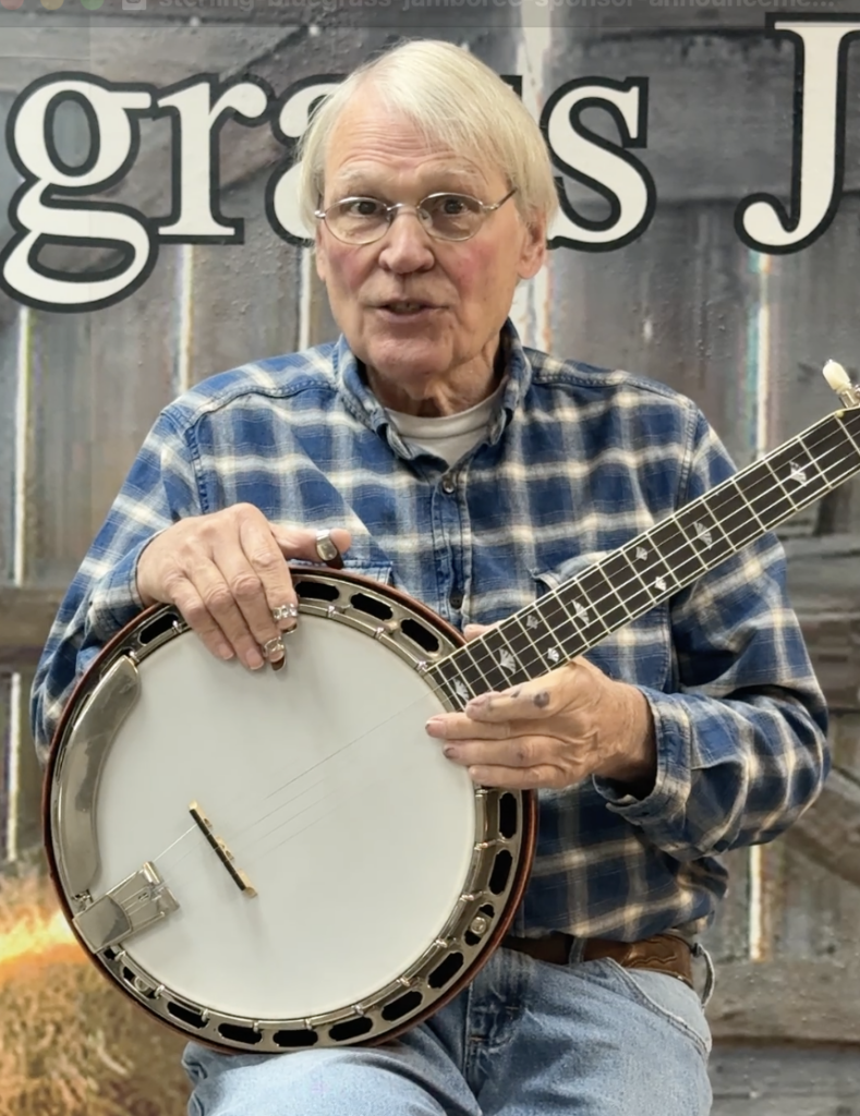 Geoff Hohwald playing banjo on stage at the Sterling Bluegrass Jamboree in Mt. Sterling, Ohio, wearing a blue plaid shirt with the venue's backdrop behind him.