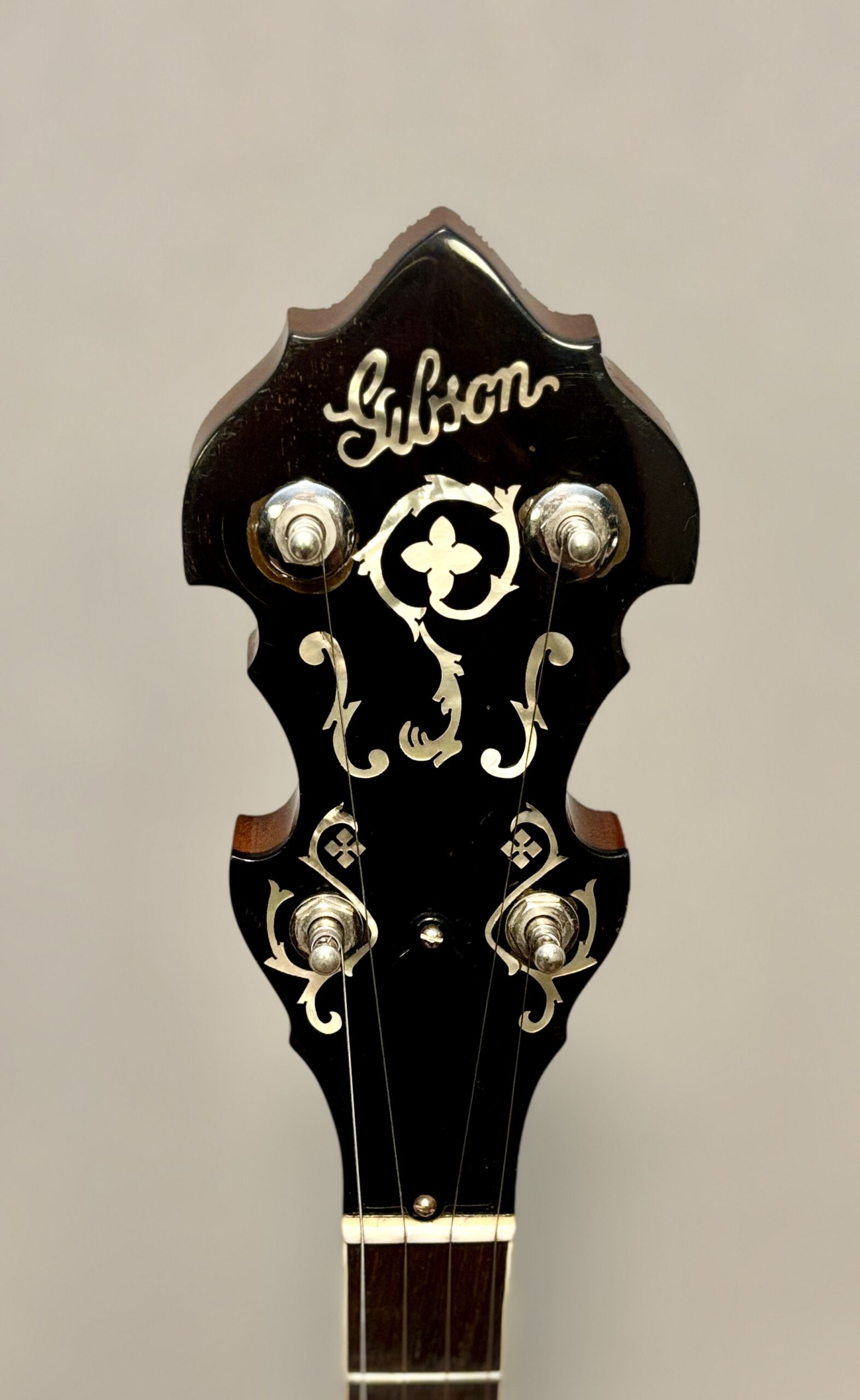 Close-up of Gibson banjo headstock featuring Gibson script logo, four-leaf clover center inlay surrounded by ornate pearl scrollwork, and nickel planetary tuners